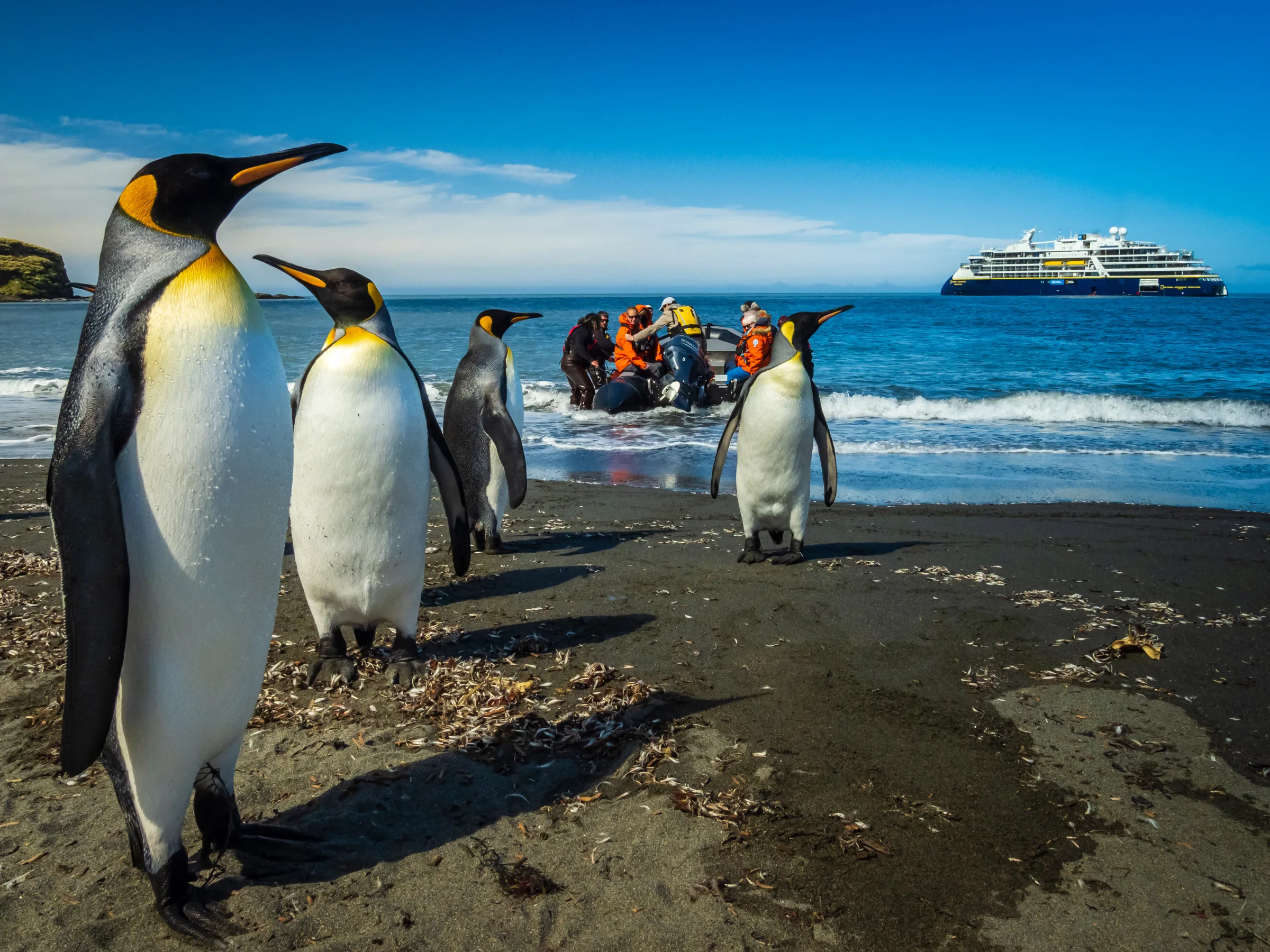 Lindblad-style expedition ship anchored in a remote icy bay at golden hour, with a zodiac of explorers in red parkas approaching shore